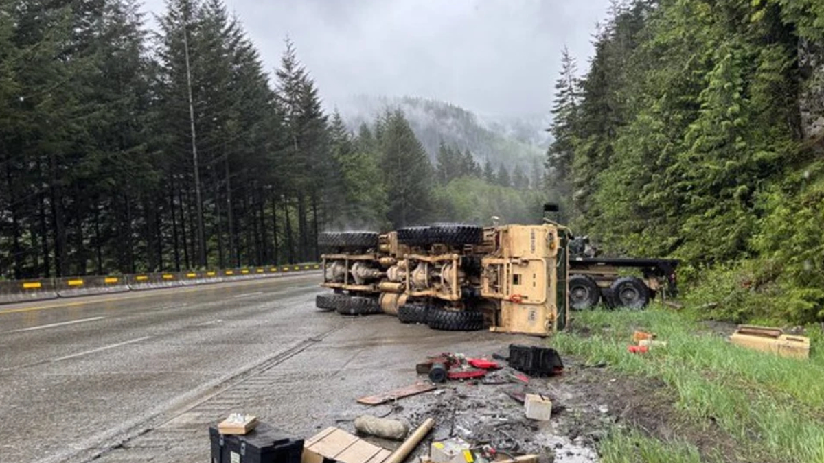 Yellow dump truck lies on its side across a wet forest highway after a crash, with scattered debris along the shoulder.