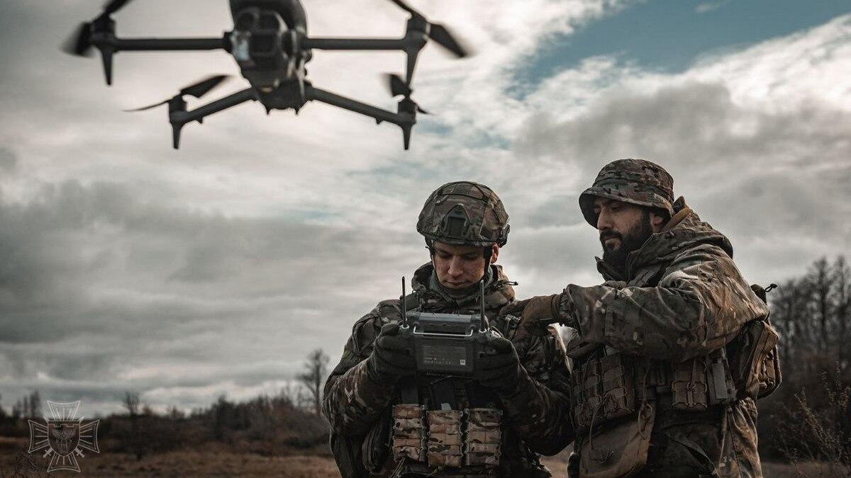 Two soldiers in camouflage gear monitor a handheld controller as a quadcopter drone flies overhead in a barren field.