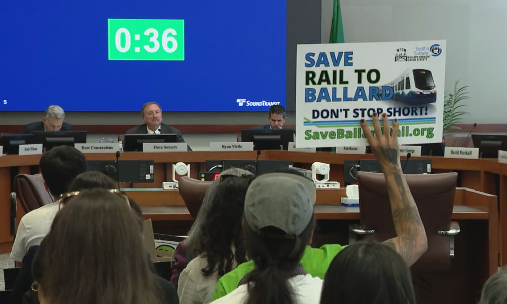 Panel of officials seated at a long desk in a hearing room; a large blue countdown screen reads 0:36 behind them while an audience member holds a ‘Save Rail to Ballard’ protest sign nearby.