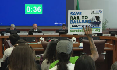 Panel of officials seated at a long desk in a hearing room; a large blue countdown screen reads 0:36 behind them while an audience member holds a ‘Save Rail to Ballard’ protest sign nearby.