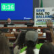 Panel of officials seated at a long desk in a hearing room; a large blue countdown screen reads 0:36 behind them while an audience member holds a ‘Save Rail to Ballard’ protest sign nearby.