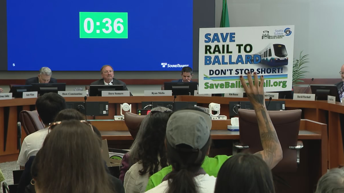 Panel of officials seated at a long desk in a hearing room; a large blue countdown screen reads 0:36 behind them while an audience member holds a ‘Save Rail to Ballard’ protest sign nearby.