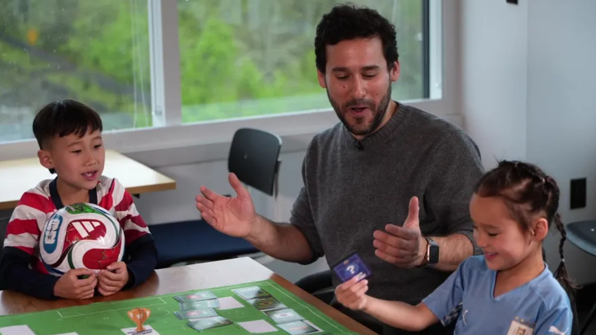 Man leads a kids' board game at a table with a rugby ball, two children smiling and holding cards by a window.