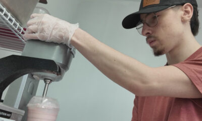 Young man in a cap and glasses wearing gloves operates a commercial mixer, pouring a pink smoothie into a cup.