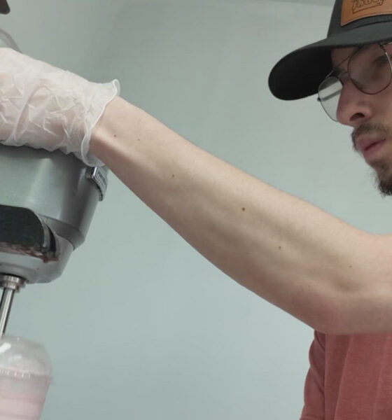Young man in a cap and glasses wearing gloves operates a commercial mixer, pouring a pink smoothie into a cup.