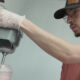 Young man in a cap and glasses wearing gloves operates a commercial mixer, pouring a pink smoothie into a cup.
