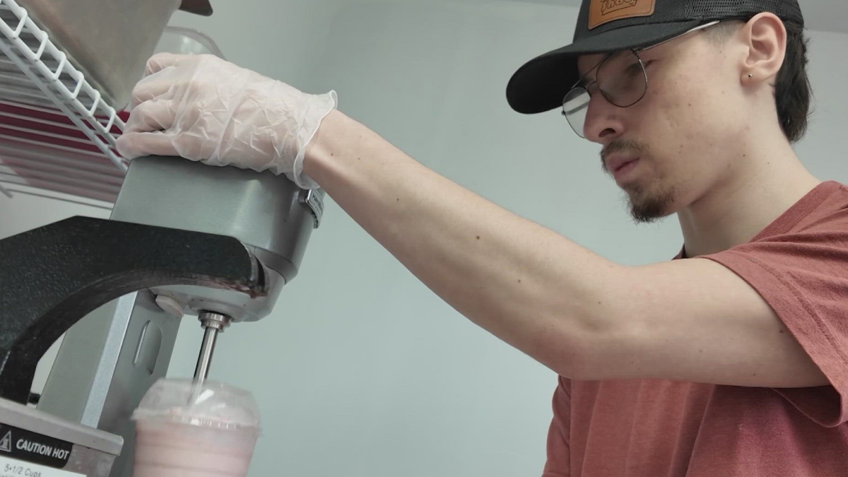 Young man in a cap and glasses wearing gloves operates a commercial mixer, pouring a pink smoothie into a cup.