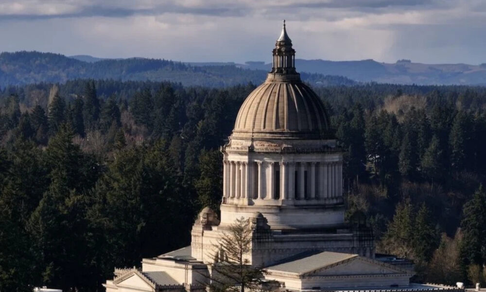 Neoclassical capitol dome rising from a stone building, surrounded by dense evergreen forest and distant hills in the background.