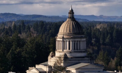 Neoclassical capitol dome rising from a stone building, surrounded by dense evergreen forest and distant hills in the background.