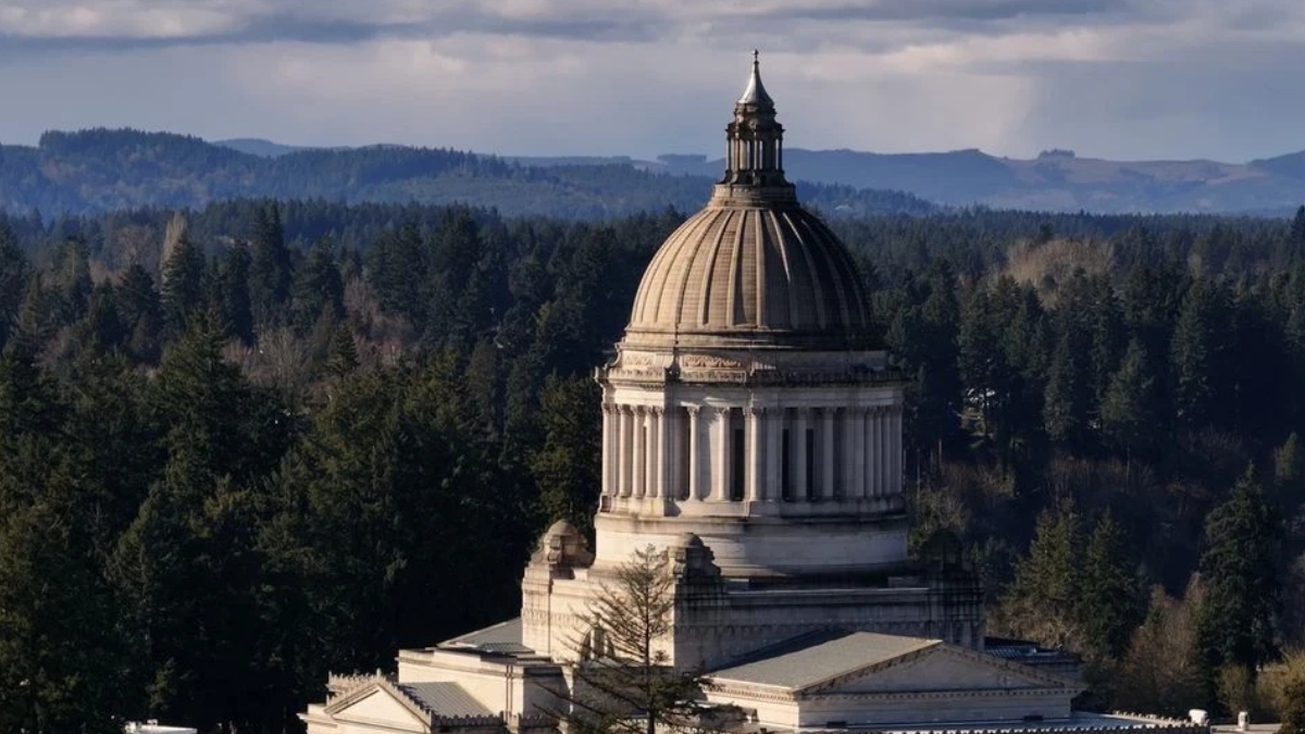 Neoclassical capitol dome rising from a stone building, surrounded by dense evergreen forest and distant hills in the background.
