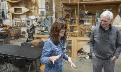 Two people chat in a bustling woodshop, a woman in denim jacket gesturing while speaking to a man in a gray pullover.