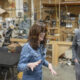 Two people chat in a bustling woodshop, a woman in denim jacket gesturing while speaking to a man in a gray pullover.