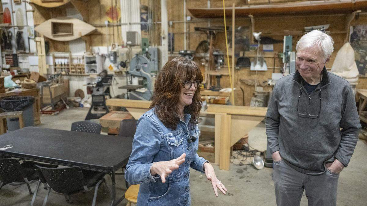 Two people chat in a bustling woodshop, a woman in denim jacket gesturing while speaking to a man in a gray pullover.