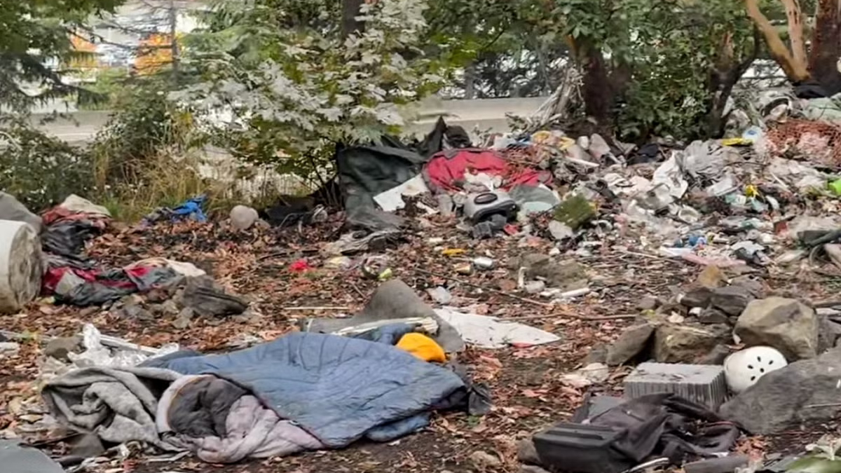 Piles of trash and discarded belongings scattered under trees, including blankets, bags, and plastic debris.