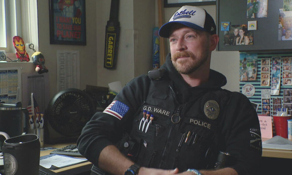 Police officer wearing a vest labeled POLICE with name G.D. Ware, seated in a cluttered office filled with photos and supplies, looking to the side.