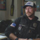 Police officer wearing a vest labeled POLICE with name G.D. Ware, seated in a cluttered office filled with photos and supplies, looking to the side.