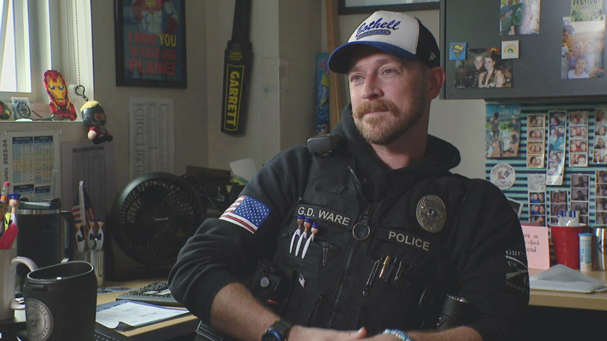 Police officer wearing a vest labeled POLICE with name G.D. Ware, seated in a cluttered office filled with photos and supplies, looking to the side.