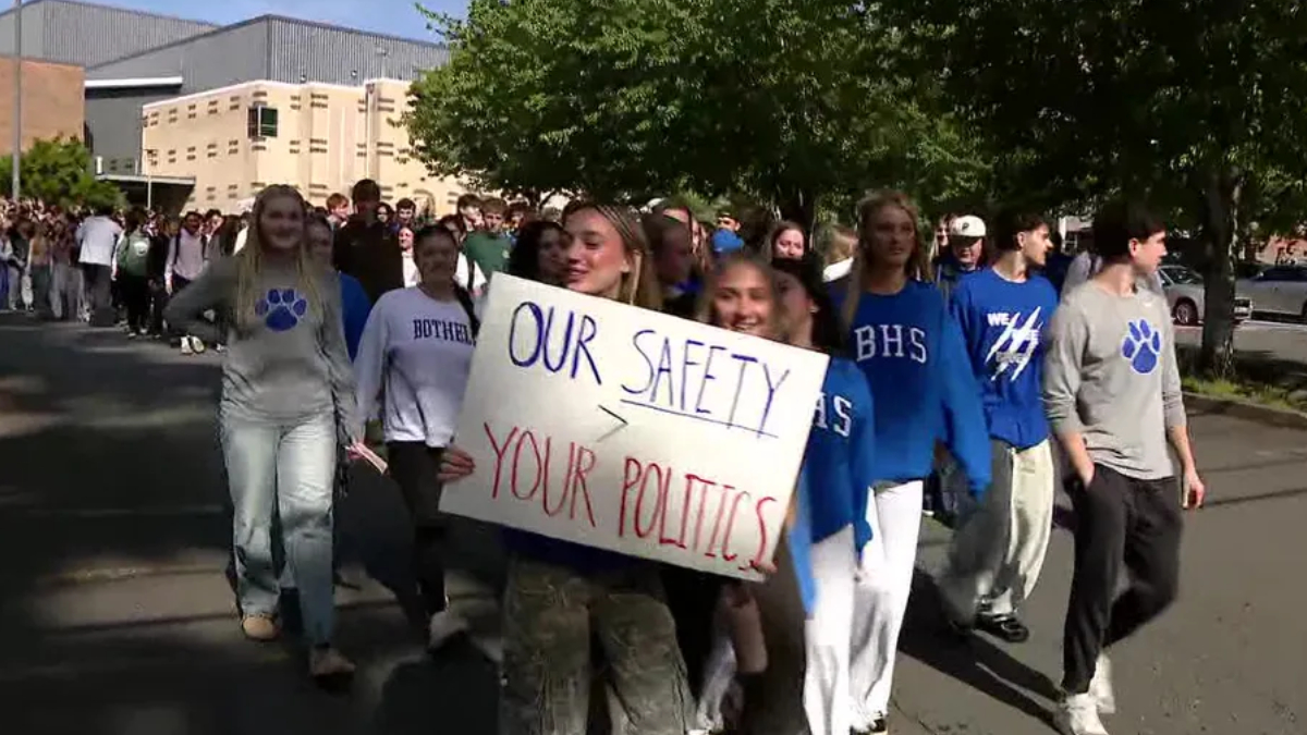 Group of students marching outside, holding a sign that reads "Our Safety > Your Politics" during a campus demonstration.