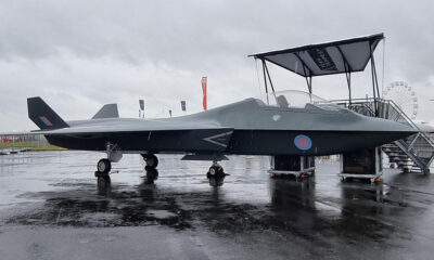 Dark gray fighter jet on a rain-soaked tarmac, RAF roundel on the fuselage, display stairs nearby on a cloudy day.