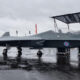 Dark gray fighter jet on a rain-soaked tarmac, RAF roundel on the fuselage, display stairs nearby on a cloudy day.