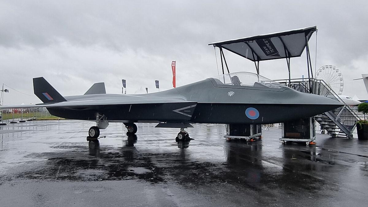 Dark gray fighter jet on a rain-soaked tarmac, RAF roundel on the fuselage, display stairs nearby on a cloudy day.