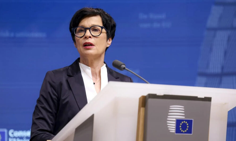 Woman in a dark blazer delivers a speech at a podium with the EU emblem against a blue backdrop.