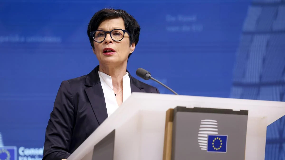 Woman in a dark blazer delivers a speech at a podium with the EU emblem against a blue backdrop.