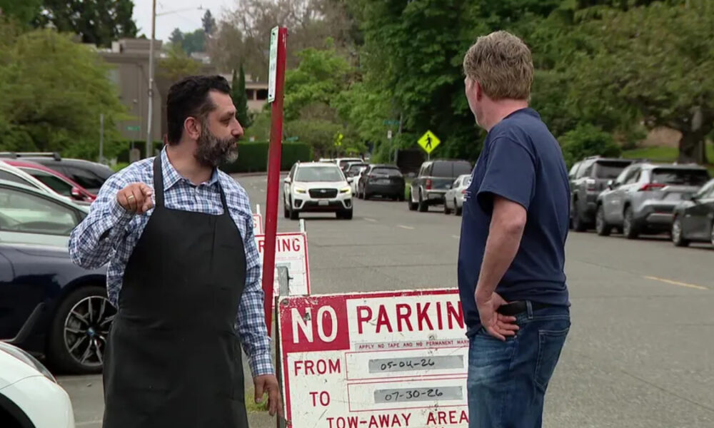 Man in a black apron points at the other man while standing beside a 'No Parking' sign in a parking lot street scene.