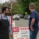 Man in a black apron points at the other man while standing beside a 'No Parking' sign in a parking lot street scene.