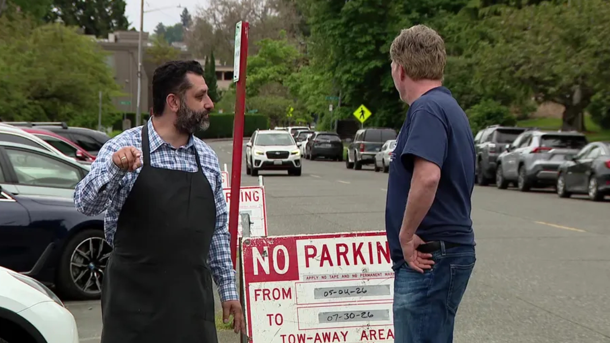 Man in a black apron points at the other man while standing beside a 'No Parking' sign in a parking lot street scene.