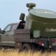 Pickup truck with a large ThinkKom industrial unit mounted on a flatbed, standing in a grassy field.