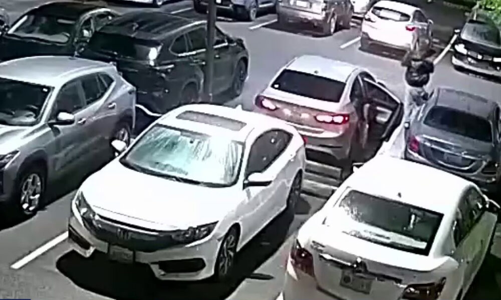 White sedan with open driver's door in a crowded parking lot; a person stands near it on the right side of the image.
