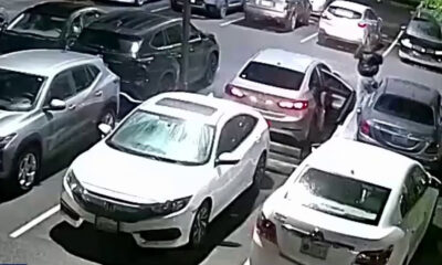 White sedan with open driver's door in a crowded parking lot; a person stands near it on the right side of the image.