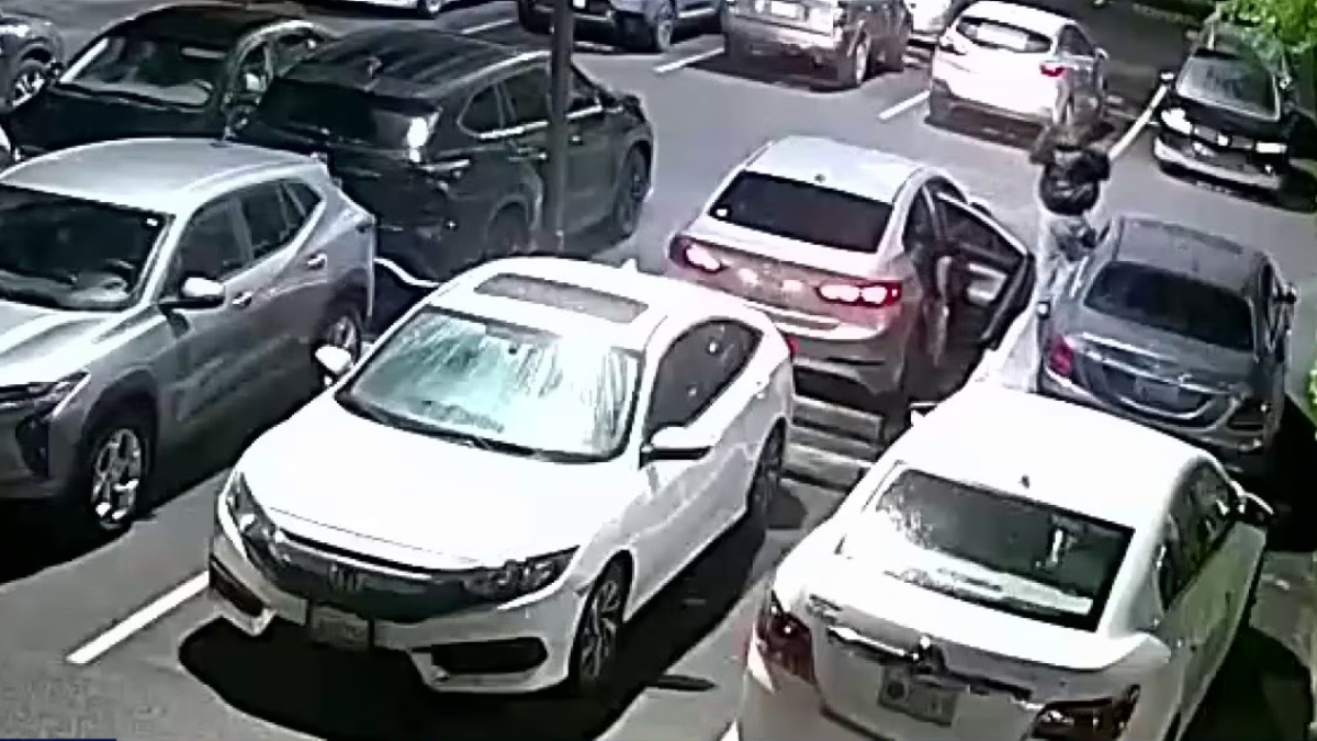 White sedan with open driver's door in a crowded parking lot; a person stands near it on the right side of the image.