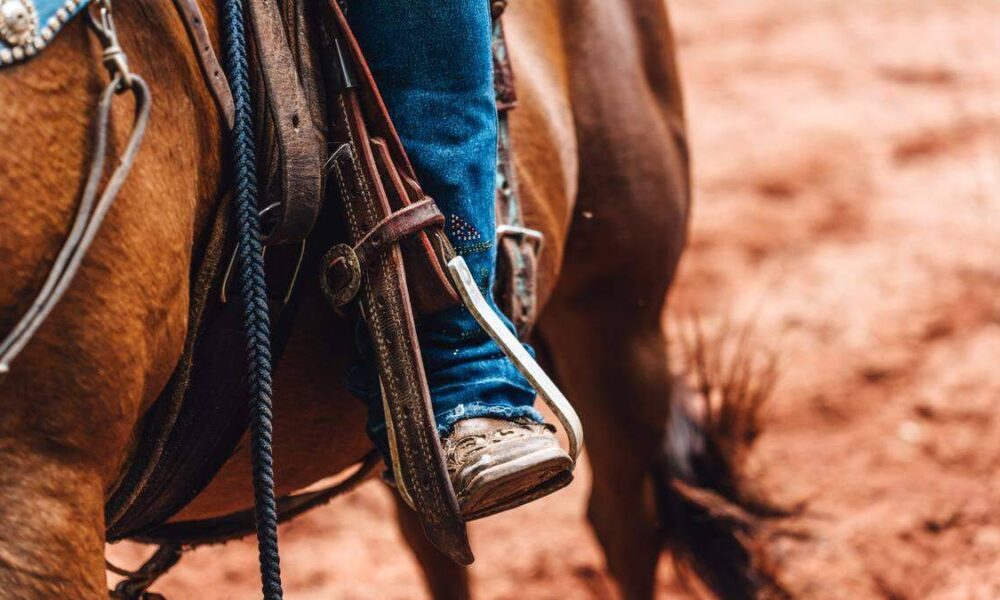 Close-up of a rider's boot in a western stirrup, strapped to a saddle on a brown horse, with dusty ground in the background.