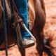 Close-up of a rider's boot in a western stirrup, strapped to a saddle on a brown horse, with dusty ground in the background.