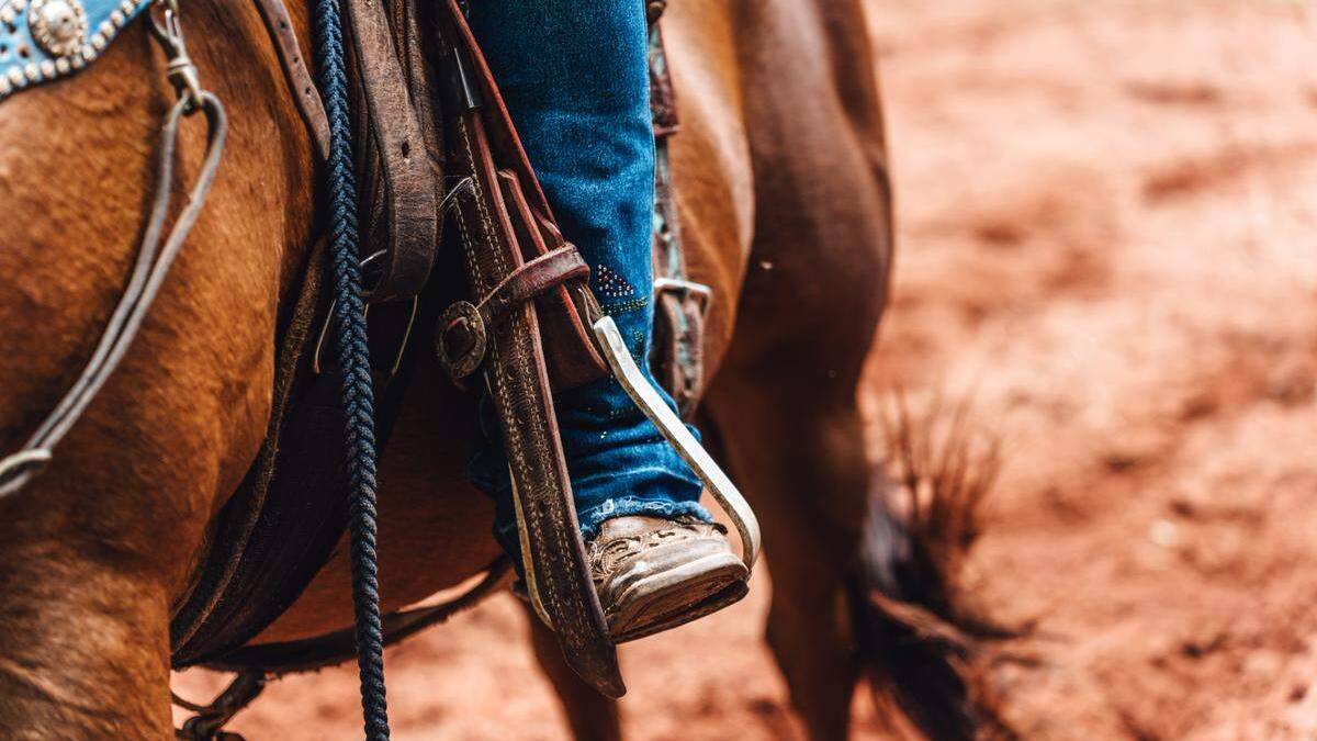 Close-up of a rider's boot in a western stirrup, strapped to a saddle on a brown horse, with dusty ground in the background.