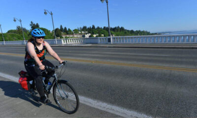 Woman cycling on a bridge wearing a blue helmet and floral tank top, with red panniers on a sunny day.