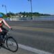 Woman cycling on a bridge wearing a blue helmet and floral tank top, with red panniers on a sunny day.