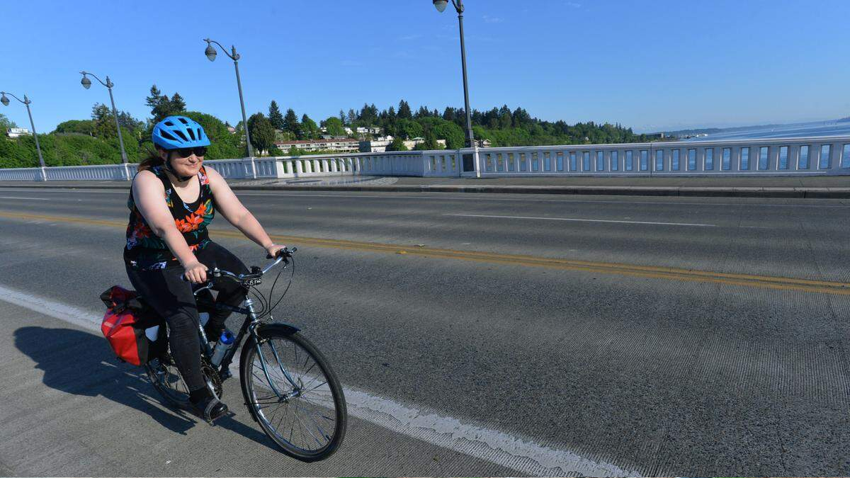 Woman cycling on a bridge wearing a blue helmet and floral tank top, with red panniers on a sunny day.