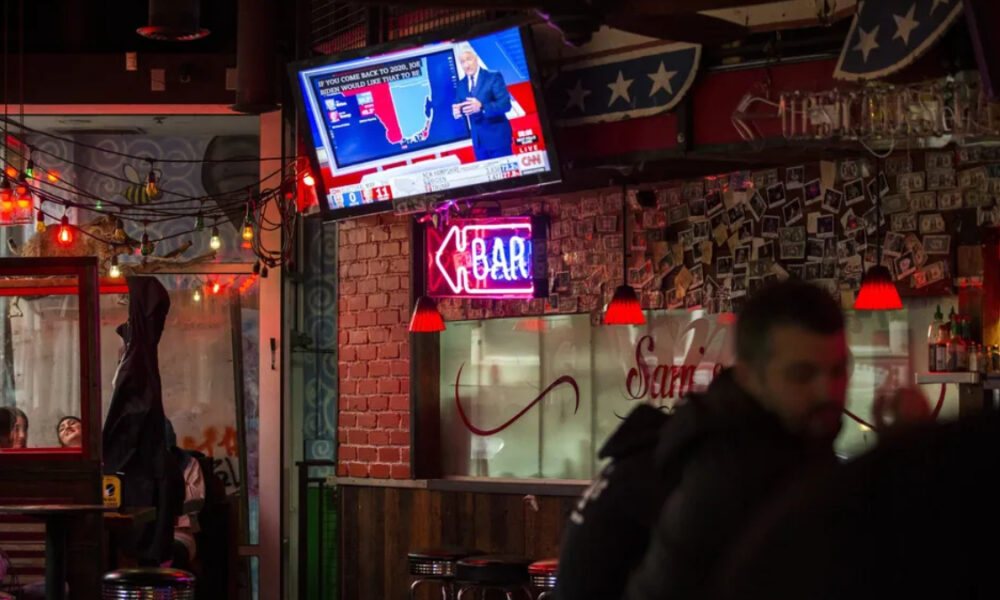 Bar interior at night with a wall-mounted TV showing a news broadcast, string lights, and a neon 'OPEN' sign above a brick pillar.