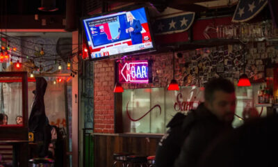 Bar interior at night with a wall-mounted TV showing a news broadcast, string lights, and a neon 'OPEN' sign above a brick pillar.
