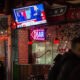 Bar interior at night with a wall-mounted TV showing a news broadcast, string lights, and a neon 'OPEN' sign above a brick pillar.
