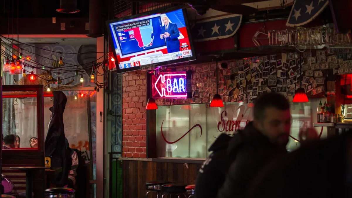 Bar interior at night with a wall-mounted TV showing a news broadcast, string lights, and a neon 'OPEN' sign above a brick pillar.