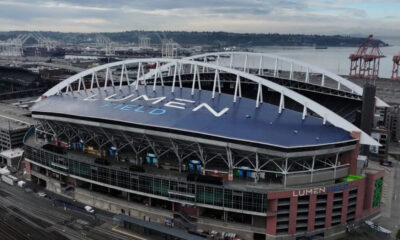 Aerial view of Lumen Field stadium with a blue roof and large white letters spelling LUMEN FIELD, overlooking a port area with cranes nearby.