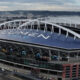 Aerial view of Lumen Field stadium with a blue roof and large white letters spelling LUMEN FIELD, overlooking a port area with cranes nearby.