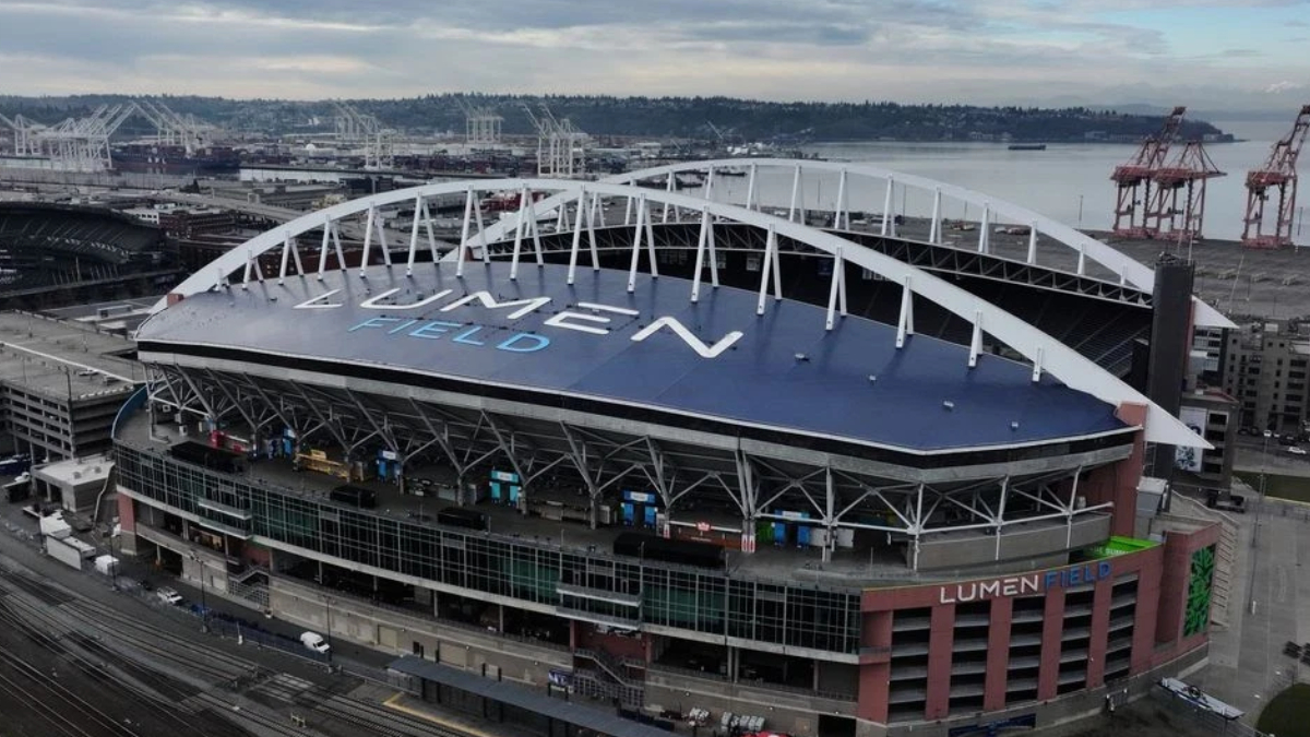 Aerial view of Lumen Field stadium with a blue roof and large white letters spelling LUMEN FIELD, overlooking a port area with cranes nearby.