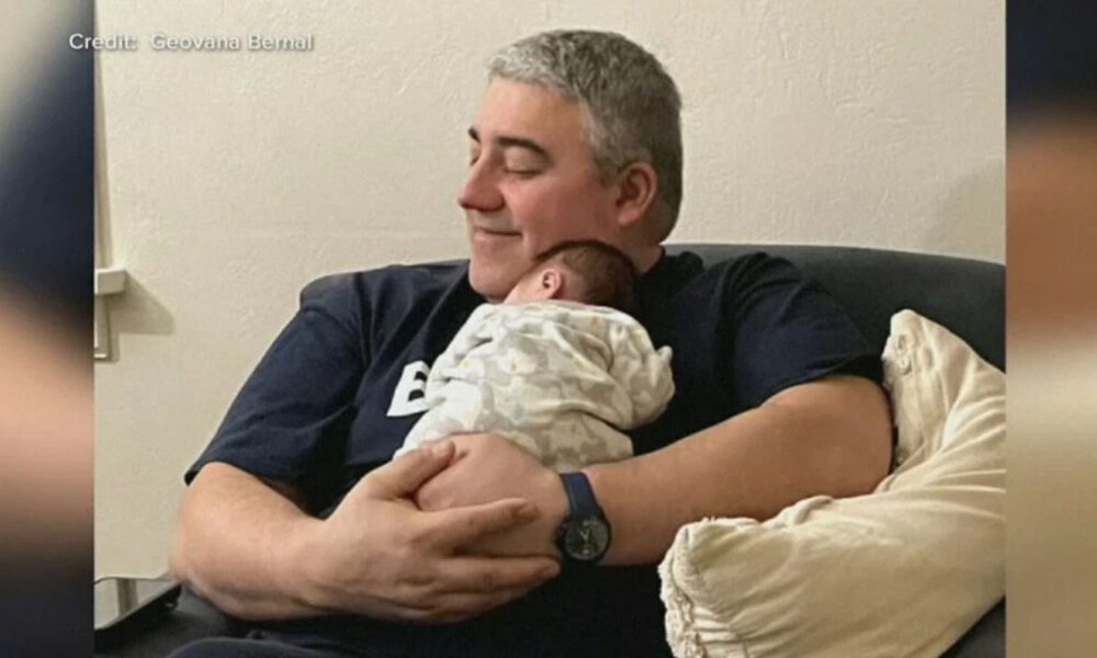 Man with gray hair cradles a newborn baby on his chest while seated on a couch, smiling softly.