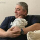 Man with gray hair cradles a newborn baby on his chest while seated on a couch, smiling softly.