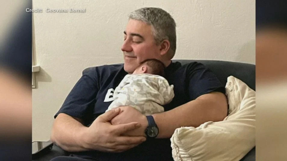 Man with gray hair cradles a newborn baby on his chest while seated on a couch, smiling softly.